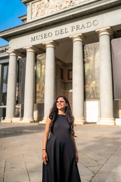 Latina happy woman in dress smiles with confidence and joy in front of Museo del Prado, Madrid, Spain. Her relaxed attitude flowing hair and sunglasses reflect a vibrant and elegant tourist in a sunny