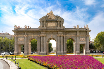 Obraz premium The Puerta de Alcala stands in Madrid Spain under a vibrant summer sky. Colorful flowers bloom in front of the historic neoclassical monument, combination of heritage, elegant city architecture