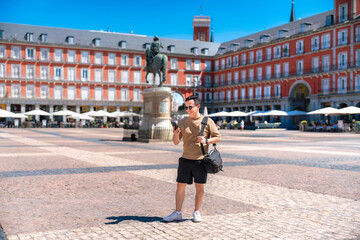 Naklejka premium Happy young Latin man smiling while checking his smartphone at Plaza Mayor in Madrid Spain, carrying a bag, wearing casual summer clothes, near the equestrian statue and iconic red historic buildings