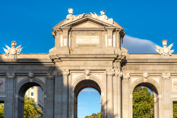 Obraz premium Puerta de Alcalá stands tall under afternoon sunlight in Madrid, Spain. The blue sky and shadows enhance its neoclassical design and sculptural details, framed by greenery at Plaza de la Independencia