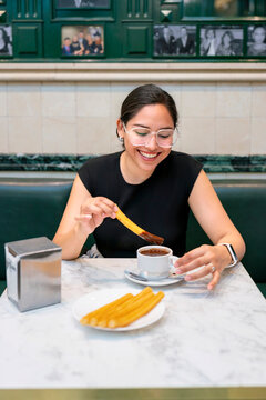 Young Latina woman smiles joyfully while dipping churros into hot chocolate at Chocolater&iacute;a San Gin&eacute;s in Madrid Spain. The famous marble tables and classic green decor highlight charm of historic cafe