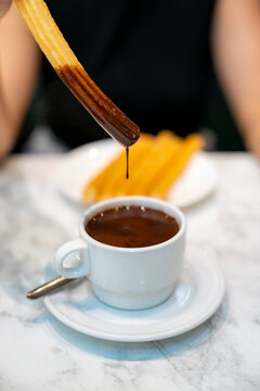 Close-up of a crispy churro dripping with thick hot chocolate inside Chocolater&iacute;a San Gin&eacute;s, an iconic and historic cafe in central Madrid, Spain, served on a marble table with classic ceramic cups
