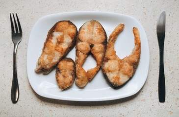 Pieces of fish fried in vegetable oil lie on a white plate, near a fork and knife. Concept for breakfast, lunch, or dinner.