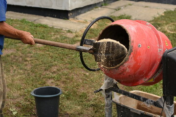 A man shovels sand into a red concrete mixer outdoors. A black bucket stands nearby. Concept of...