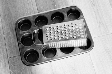 A monochrome still life composition of essential kitchen utensils, featuring a metal box grater placed on a twelve-cup muffin baking tin on a wooden surface