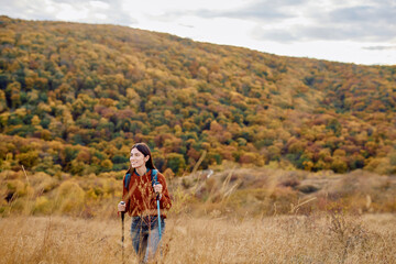 Woman with backpack and trekking poles enjoying an autumn hike, walking through dry grass with a...