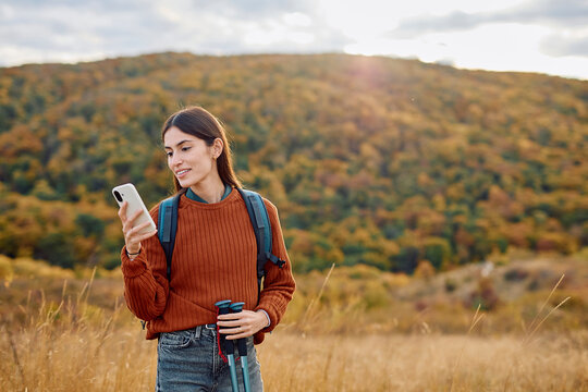 Woman standing on a hill checking her smartphone while hiking, wearing a backpack and holding trekking poles in autumn nature