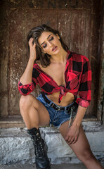 Young brunette woman with rebellious attitude sits on rustic stone steps, wearing a tied checkered shirt, denim shorts, and boots. Vintage textures and direct gaze evoke bold, rural charm.