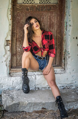 Young brunette woman with rebellious attitude sits on rustic stone steps, wearing a tied checkered shirt, denim shorts, and boots. Vintage textures and direct gaze evoke bold, rural charm.