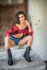 Young brunette woman with rebellious attitude sits on rustic stone steps, wearing a tied checkered shirt, denim shorts, and boots. Vintage textures and direct gaze evoke bold, rural charm.