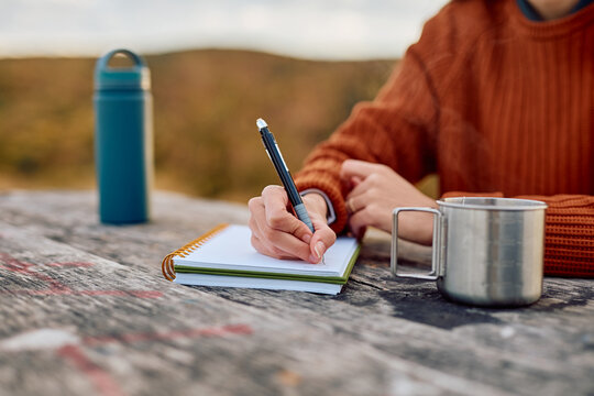 Person's hand writing in a spiral notebook, journaling thoughts on a rustic wooden table with a traveling mug and water bottle