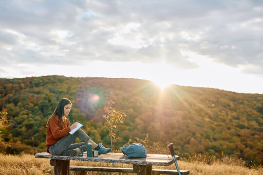 Young woman journaling on a picnic table amidst autumn foliage during golden hour, finding inspiration in nature
