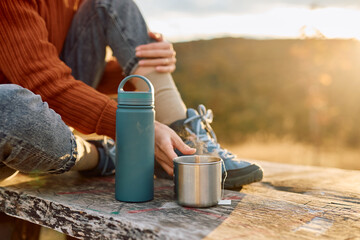 Woman sitting on a wooden surface, holding a hot steamy metal mug while a water bottle sits nearby, basking in the warm glow of sunset