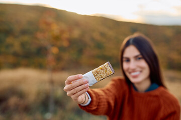 Woman smiling, holding out a granola bar, enjoying a healthy snack during a hike in an autumnal...