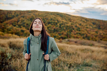 Young woman standing on a hill, eyes closed, breathing fresh air and feeling serene during an autumn hike in the mountains