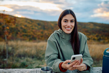 Young woman smiling, holding smartphone, relaxing outdoors during autumn hiking trip, enjoying connection and peace