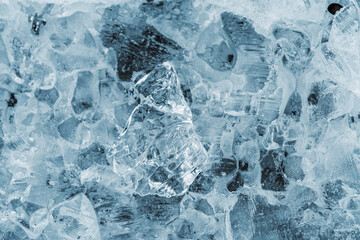Macro photograph of naturally shaped ice crystals showing intricate textures and transparency. The clear frozen surface reflects soft light and reveals layers of frozen patterns, capturing the beauty 