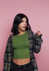 Young woman in casual attire pointing upwards against a pink background.