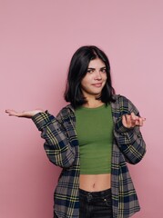 Young woman with a puzzled expression, wearing a green top and plaid shirt, standing against a pink...