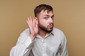Man in white shirt listening with hand to ear on beige background.