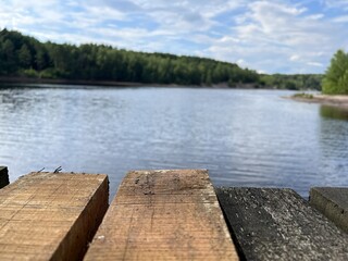 Fototapeta premium Wooden pier by a calm river on a sunny day. Tranquil nature landscape with water, forest, and blue sky. Perfect for concepts of relaxation, travel, and summer outdoors.