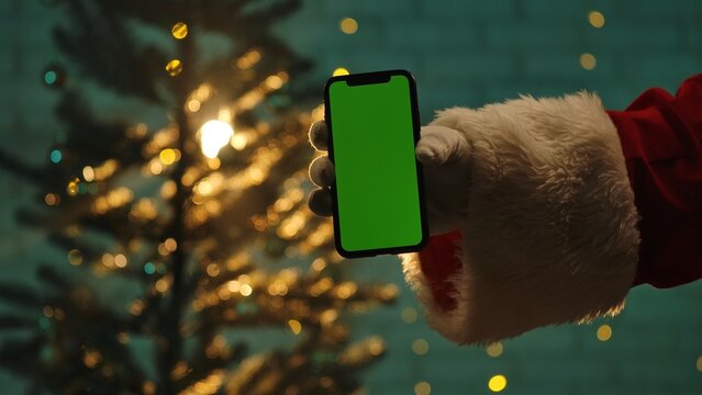 Santa hand holds a smartphone with a green screen in front of a Christmas tree, ready for customization.