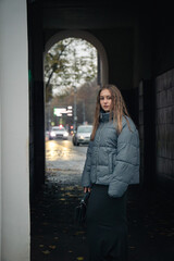 Young woman in a gray puffer jacket and black skirt walks through an urban archway on a cloudy autumn day