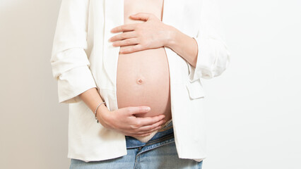 A pregnant woman stands with her hands on her rounded belly, dressed in a white blazer and blue...