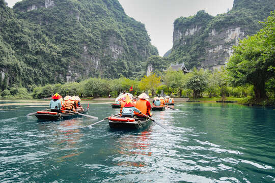 Tourists in traditional conical hats and life jackets enjoying a boat tour down the calm ngo dong river, surrounded by towering limestone karsts and green vegetation in ninh binh, vietnam