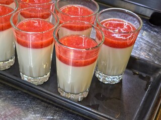 Clear glasses filled with a two-layered dessert, consisting of a creamy white base (likely panna cotta or pudding) topped with a thick, vibrant red fruit purée (likely strawberry), arranged on a tray.
