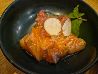 Close-up of a dish featuring cubes of raw, orange salmon marinated in a spicy or citrus dressing, garnished with thin slices of garlic and a sprig of fresh mint, served in a deep, dark bowl.