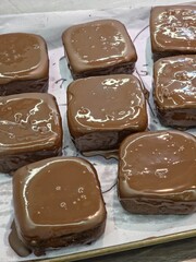 Overhead shot of approximately nine square-shaped chocolate cakes or brownies neatly arranged on a tray with baking paper, each covered in a generous, thick, and glossy coat of milk chocolate frosting