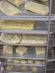 Vertical close-up of a metal cooling rack or shelf unit displaying several types of raw pastry or bread dough in various shapes (rolls, blocks, and sheeted squares), ready for baking.