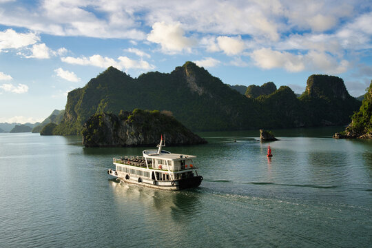 Tourist boat sailing on the calm sea of ha long bay, with towering limestone karsts emerging from the water under clear blue sky, creating a famous travel destination