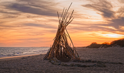Tall wooden sticks form a teepee-like structure on a quiet beach at sunset.