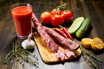 raw sausages and sliced pieces arranged on wooden cutting board, accompanied by a glass of fresh tomato juice, ripe tomatoes on the vine, cucumbers, corn, garlic, red chili peppers