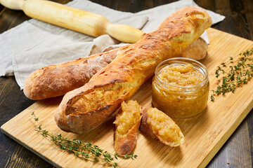Fresh baguettes with apple or quince jam on wooden board. Sliced bread spread with sweet preserve, garnished with thyme. Rolling pin and linen napkin in background. 