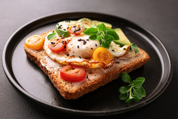Tasty homemade toast for breakfast with fried eggs, cream cheese, avocado, cherry tomatoes and fresh oregano leaves on black stone background close up.