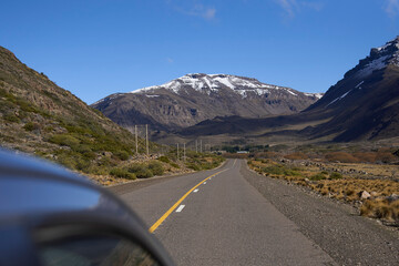 Fototapeta premium Traveling by car through Neuquen Province, Argentine Patagonia: the vehicle, the empty highway, low-lying plants along the side, and imposing snowcapped mountains in the background.