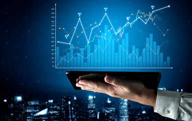 A hand holds a tablet displaying digital financial graphs and data analytics, set against a dynamic...