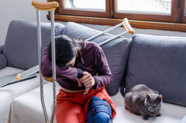 Depressed disabled young woman with her head resting on a crutch sitting on a couch.Sad woman feeling pain with her cat besides her.