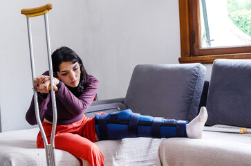 Sad young woman with crutches with a broken leg sitting on a couch.
