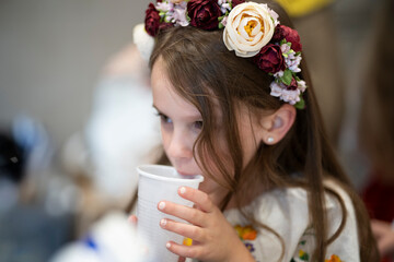 Little girl in an embroidered blouse with a floral wreath on her head drinking from a cup indoors with a blurred background, concept of thirst and childhood simplicity
