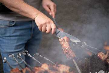 Man checking the readiness of barbecue meat with a knife on a grill, smoke rising around, concept of outdoor cooking and summer leisure.