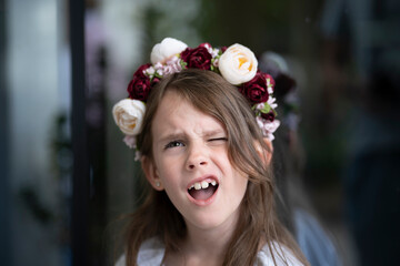 Playful little girl in a floral wreath and white blouse with her mouth open and one eye squinted, standing against a glass surface background, concept of childhood joy and spontaneity.