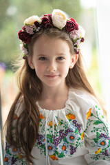 Vertical photo of a smiling Ukrainian girl in a floral wreath and white embroidered blouse standing by a window with a blurred background, concept of childhood and Ukrainian tradition.