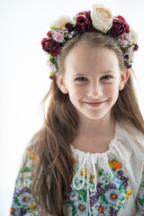 Vertical photo of a smiling Ukrainian girl in a floral wreath and white embroidered blouse against a white background with copy space, concept of childhood and Ukrainian tradition.