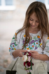 Vertical waist-up photo of a smiling Ukrainian girl in an embroidered blouse looking down at a floral wreath in her hands, concept of childhood joy and Ukrainian tradition.
