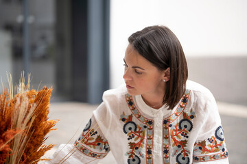 Dark-haired beautiful focused woman in an embroidered blouse creating a composition from fluffy plants outdoors, concept of creativity and do-it-yourself art