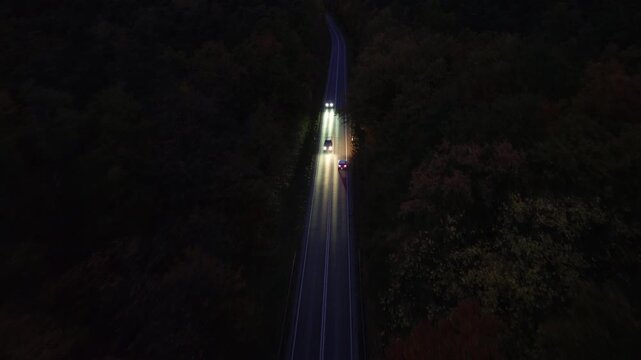 Aerial view of vehicles traveling along winding road at night, illuminated headlights cutting through darkness, showcasing dynamic motion and depth, camera follows action with smooth tracking shot